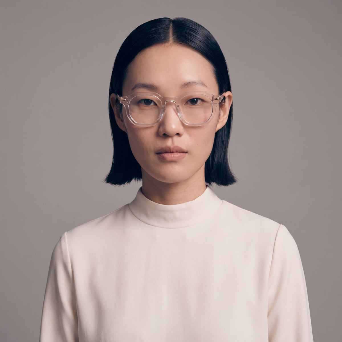 A model with short black hair wearing clear, thick-rimmed square eyeglasses and a cream-colored mock neck top, photographed against a solid neutral gray background.