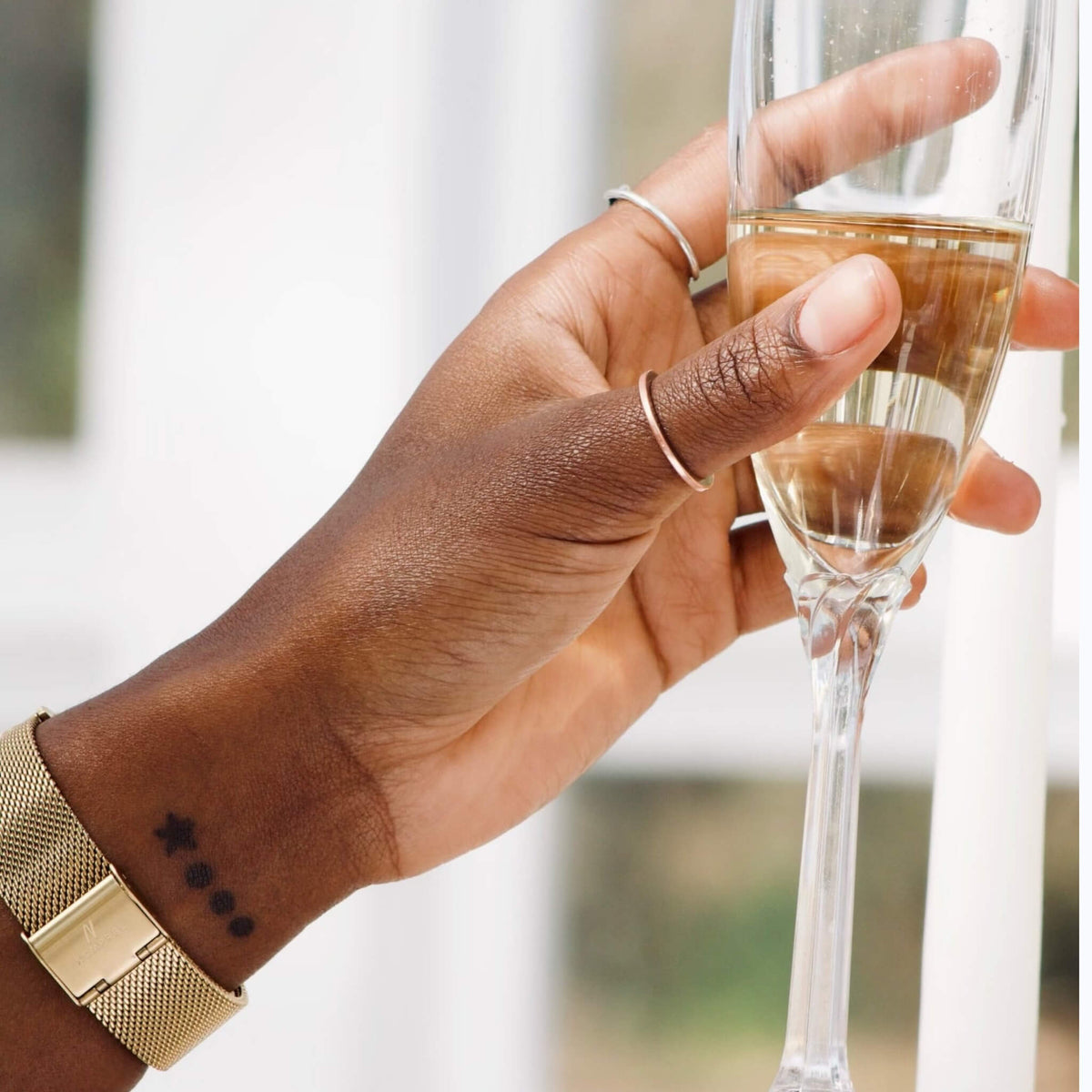 A close-up of a person's hand holding a glass of champagne, wearing a gold mesh advance champagne 36mm NCMPRBL watch