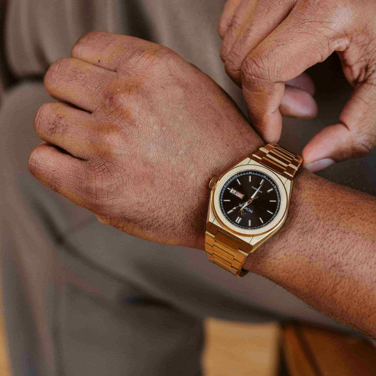 A close-up lifestyle shot of a gold-tone wristwatch with a black dial being worn on a person's wrist, with their other hand adjusting or touching the strap.