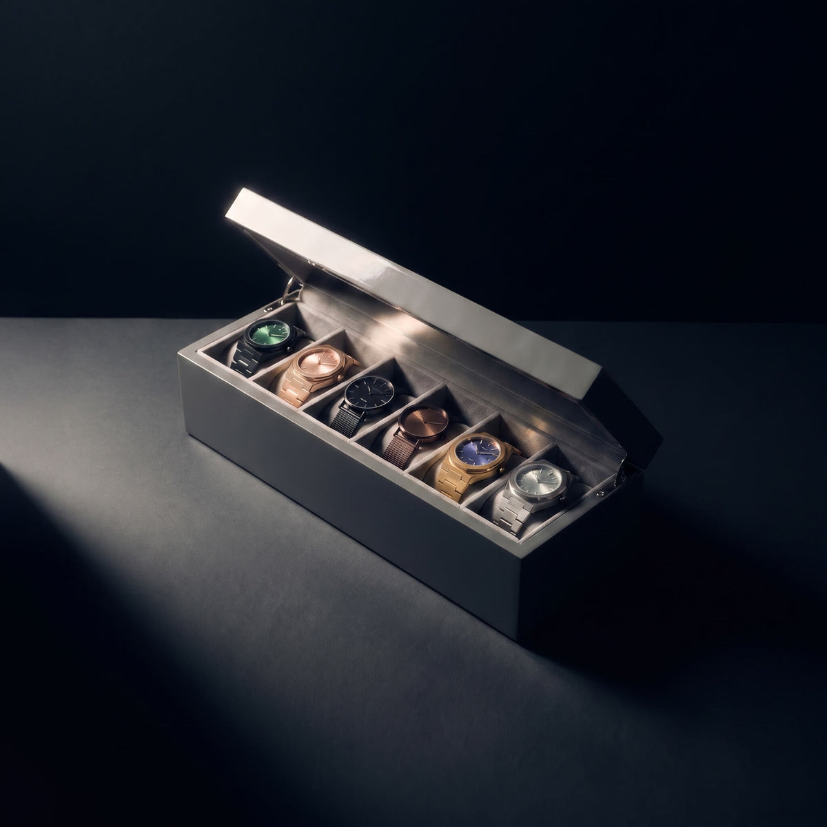 A luxury watch display box with an open lid, showcasing six different wristwatches in individual slots. The box is a dark gray wood with a brushed silver metal lid, set against a dark background with dramatic spotlighting.