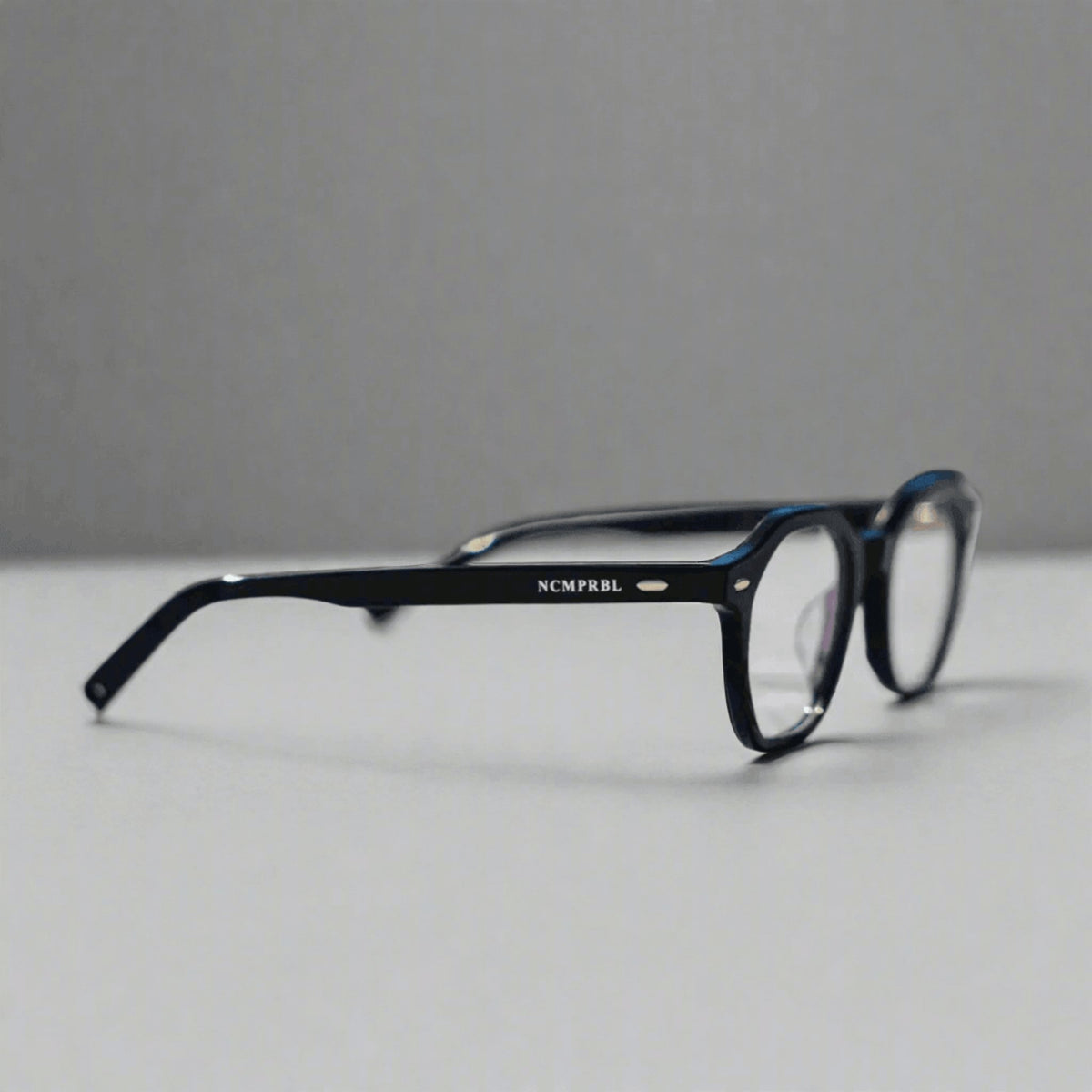 Black acetate eyeglasses with a small silver logo reading 'NCMPRBL' on the temple arm, resting on a light gray surface against a darker gray background.