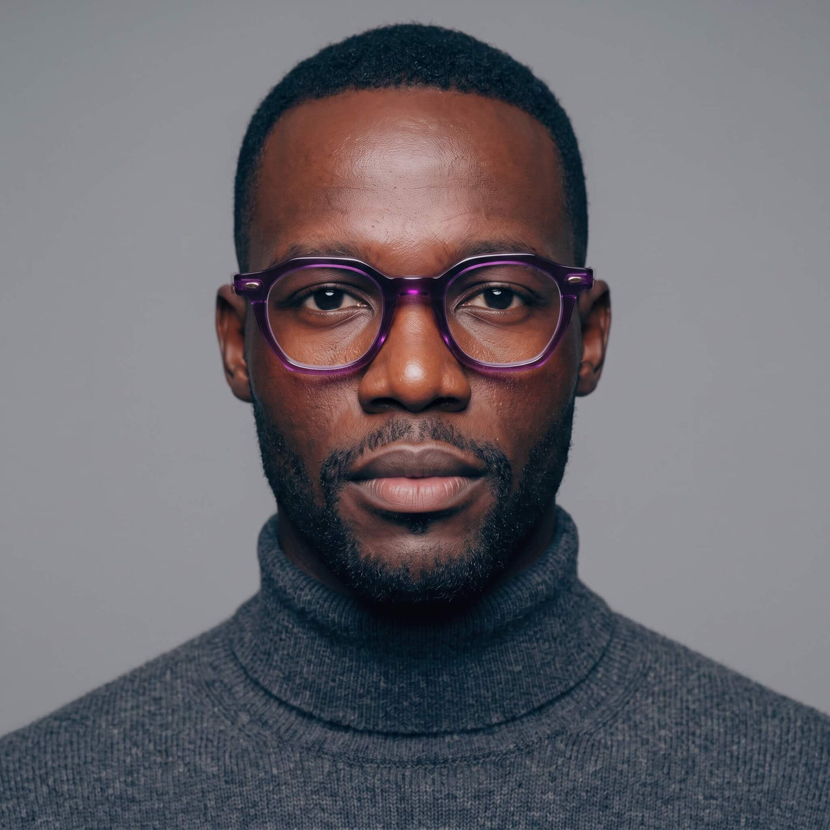 A close-up portrait of a man with short dark hair, a trimmed beard, and haze 46mm geometric purple-framed eyeglasses, wearing a dark gray turtleneck sweater against a plain gray background.