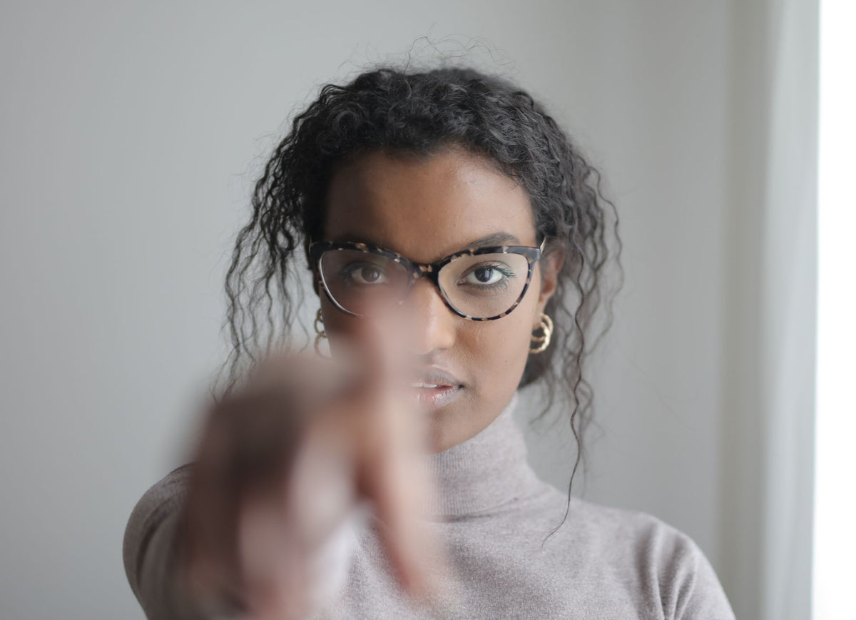 Confident African American female pointing with finger at camera looking seriously at camera through glasses on gray wall