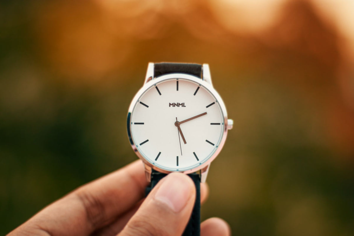 Close-up of a minimalist watch with a leather strap held by a hand against a blurred background.