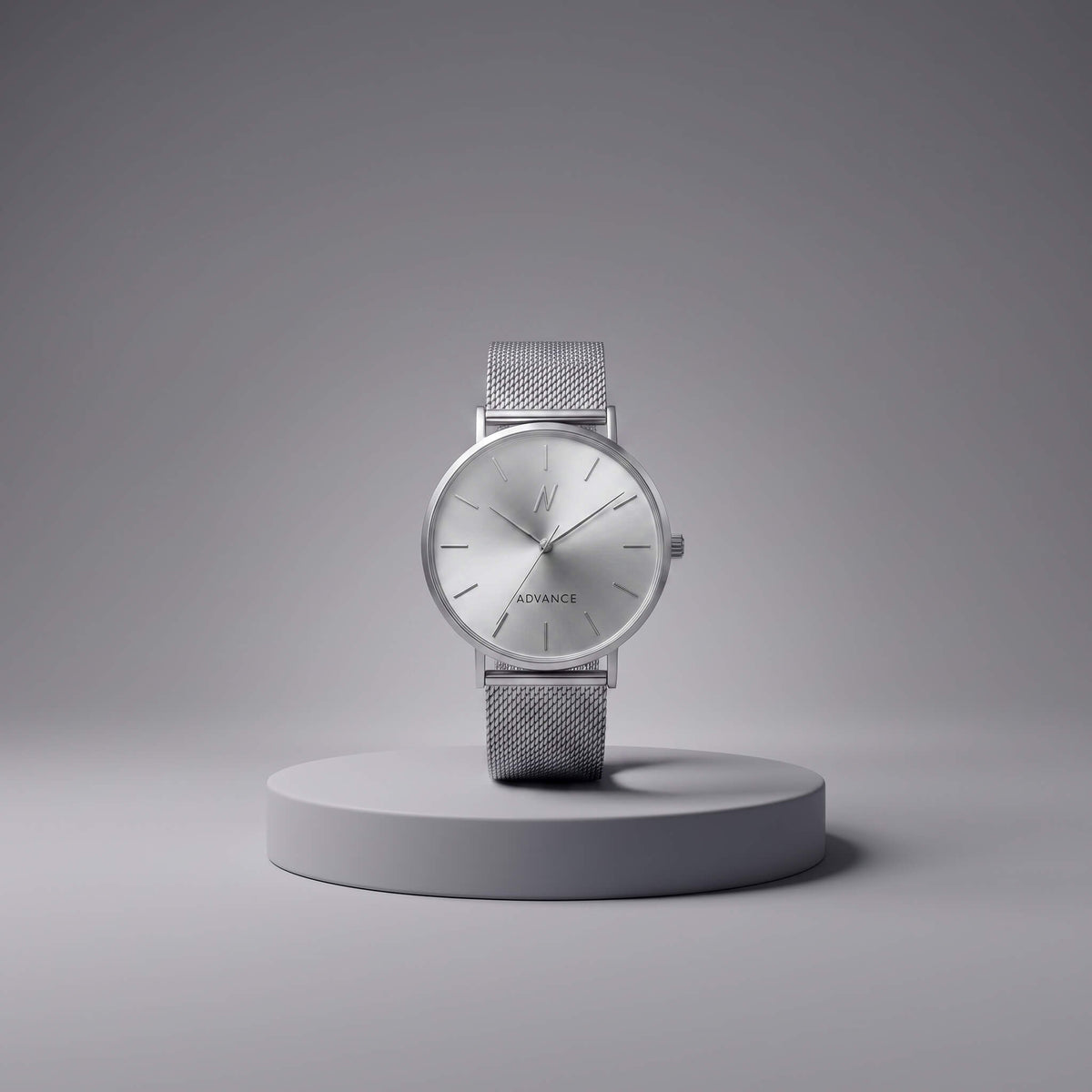 A minimalist silver analog watch with a mesh strap and a silver dial, displayed on a round gray pedestal against a clean gray background. The brand name 'ADVANCE' is visible on the dial.