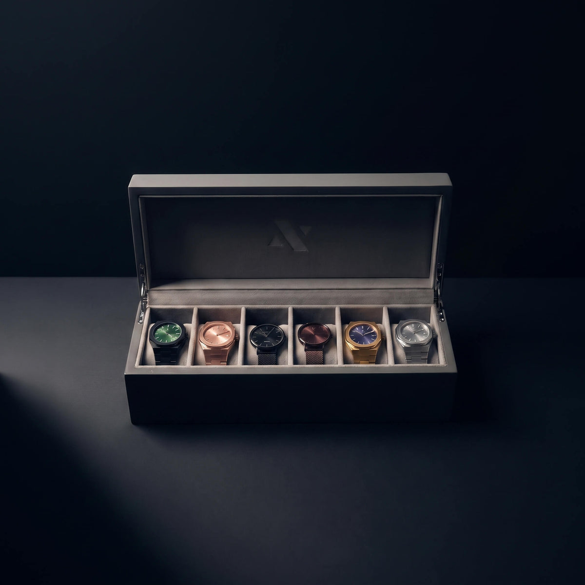 A luxury watch display box with an open lid, showcasing six different wristwatches in individual slots. The watches feature various metal cases (green, rose gold, black, copper, gold, and silver) and dials.