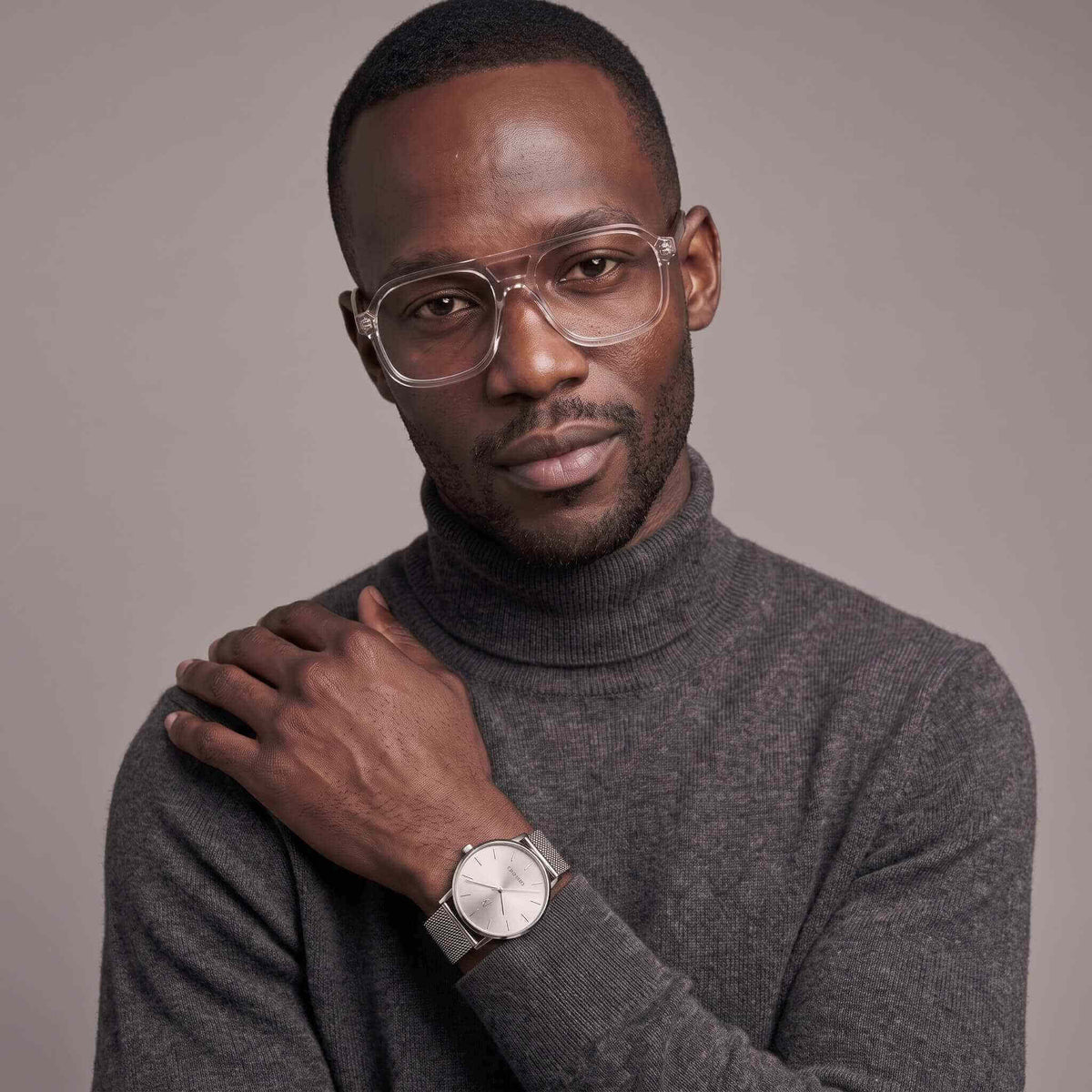 A man wearing clear-framed eyeglasses, a dark gray turtleneck sweater, and a silver watch, posed against a solid gray studio background.