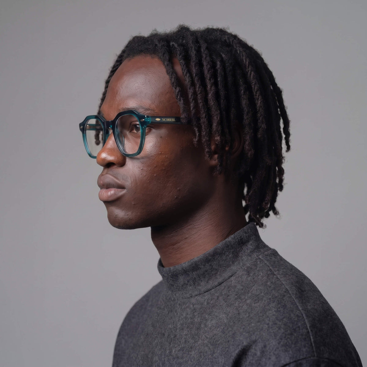 A man in profile wearing manifest geometric  eyeglasses with a translucent deep evergreen teal frame and a dark gray turtleneck sweater, set against a solid gray studio background.