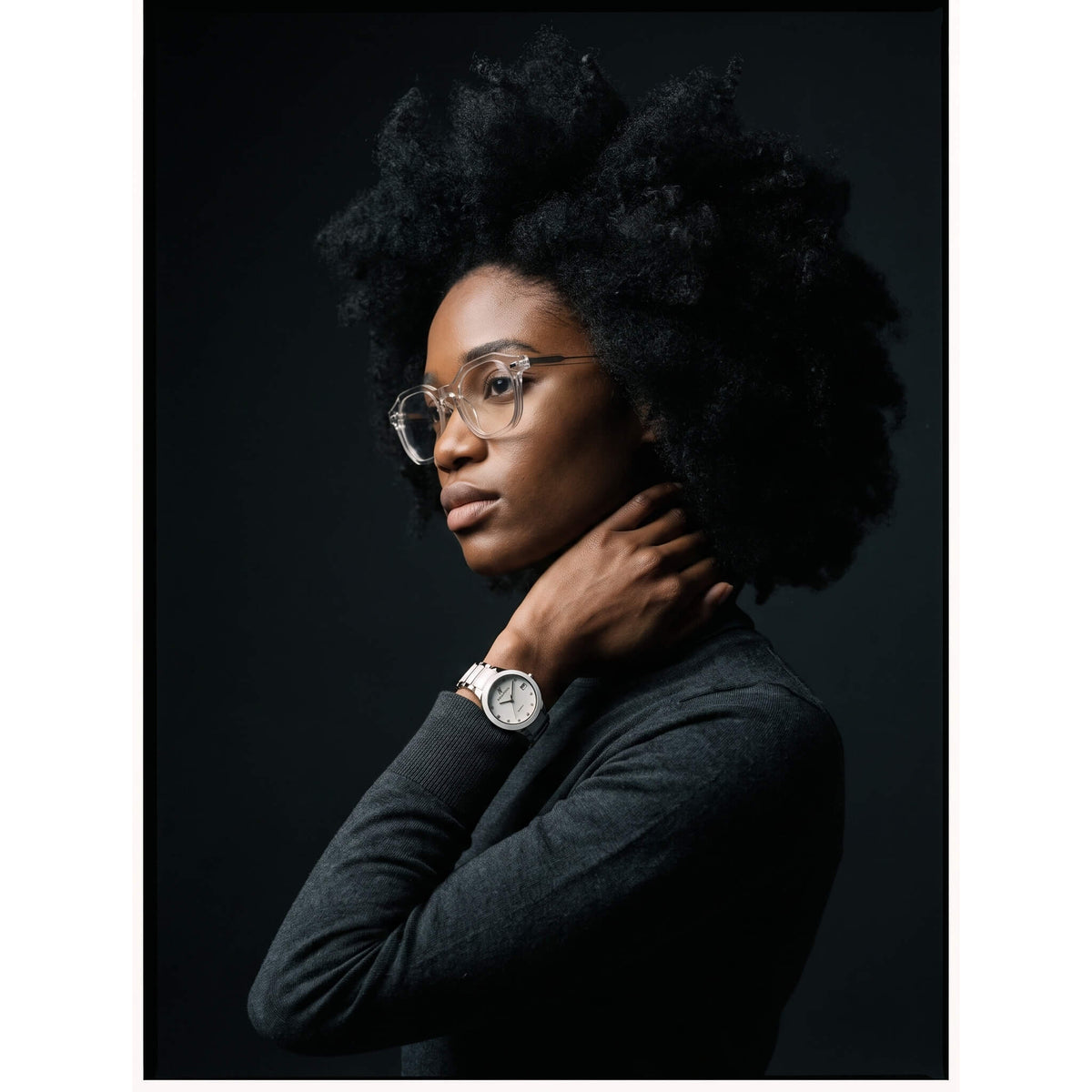 A woman with an afro hairstyle, wearing round eyeglasses, a dark gray long-sleeve top, and a silver watch with a white face, posed against a dark studio background.