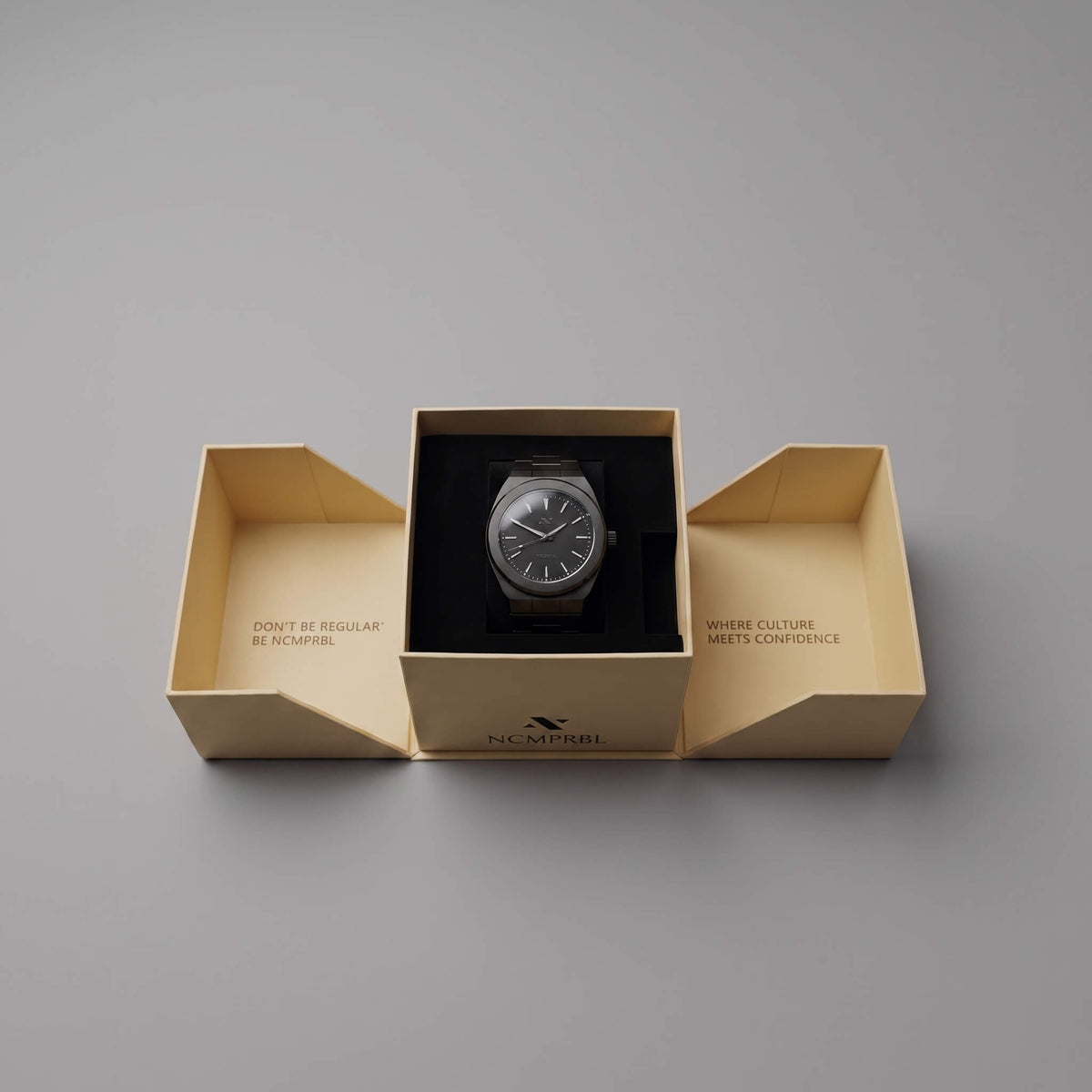 A elevate link phantom wristwatch presented in an open cream-colored branded box on a smooth gray background. The watch has a dark dial and a dark strap, centered inside a black insert, with the brand name NCMPRBL printed on the front of the box.