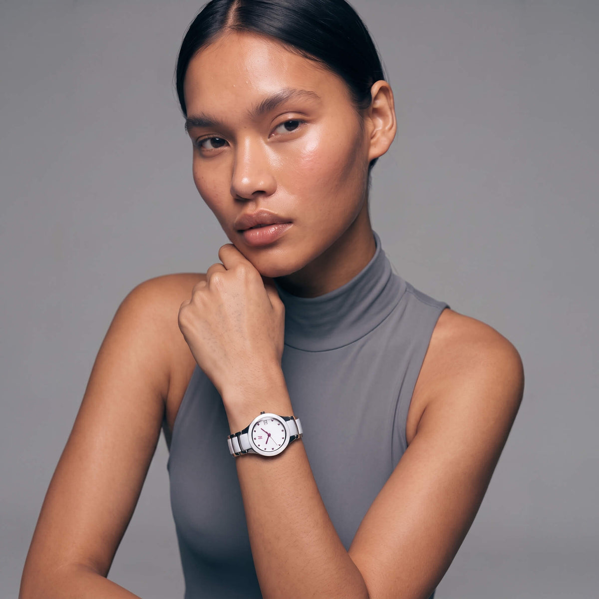 A woman wearing a minimalist silver-toned watch with a white dial and a gray sleeveless mock-neck top, posing against a solid gray studio background.